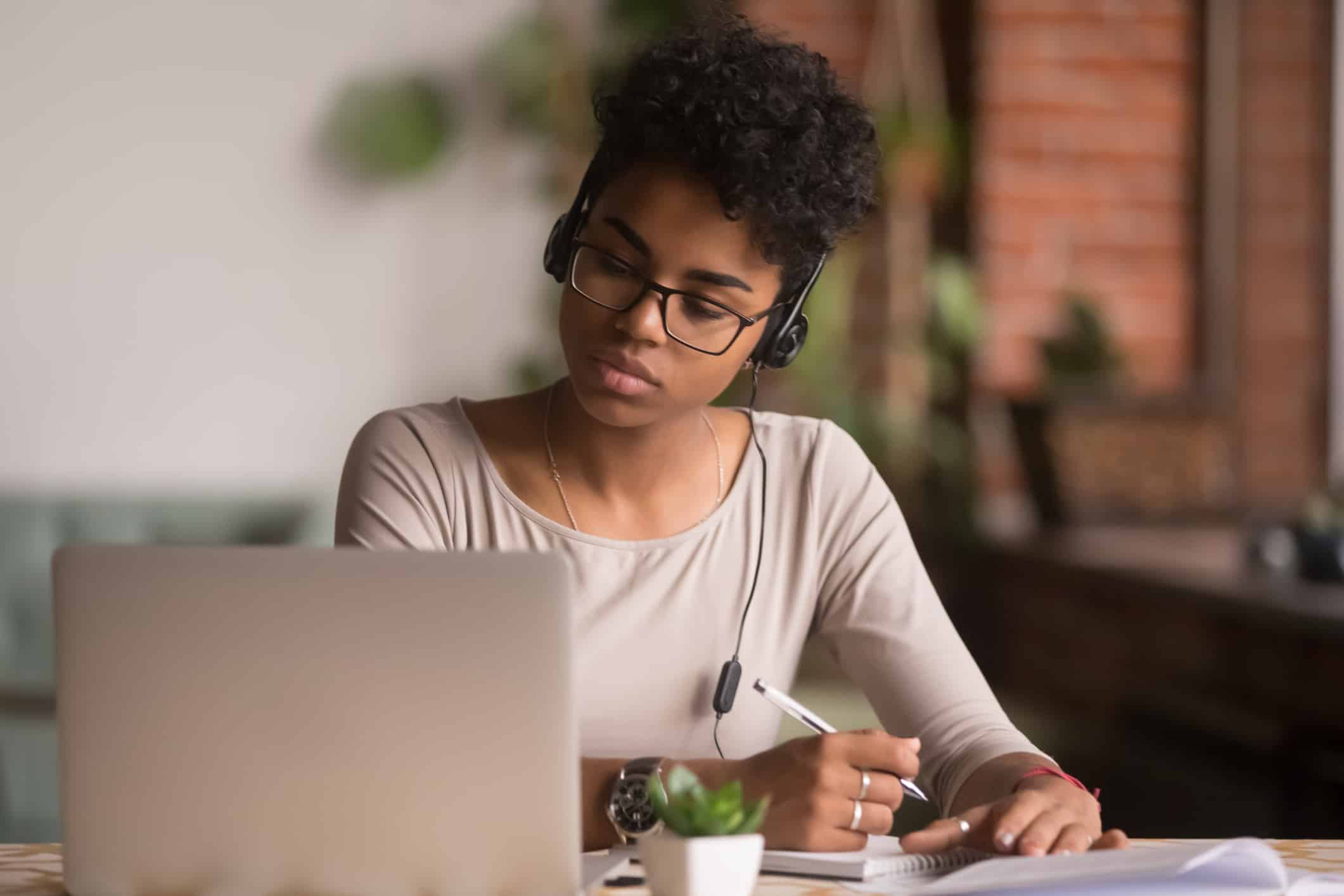 Focused mixed race woman wearing headphones watching webinar write notes Focused mixed race woman wearing headphones watching webinar write notes study online with skype teacher, african female student learning language computer course on laptop listen translate lecture