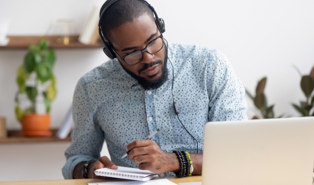 African American man with headphones on listening to a podcast on a laptop and taking notes in a notebook.