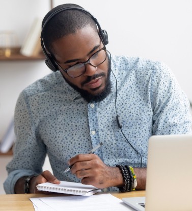 Focused african business man in headphones writing notes in notebook watching webinar video course, serious black male student looking at laptop listening lecture study online on computer e learning (Focused african business man in headphones writing