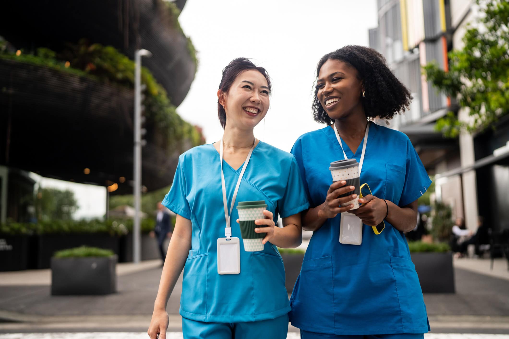 Two women in medical uniforms stroll through the city with takeaway coffee, sharing a light moment during their break.