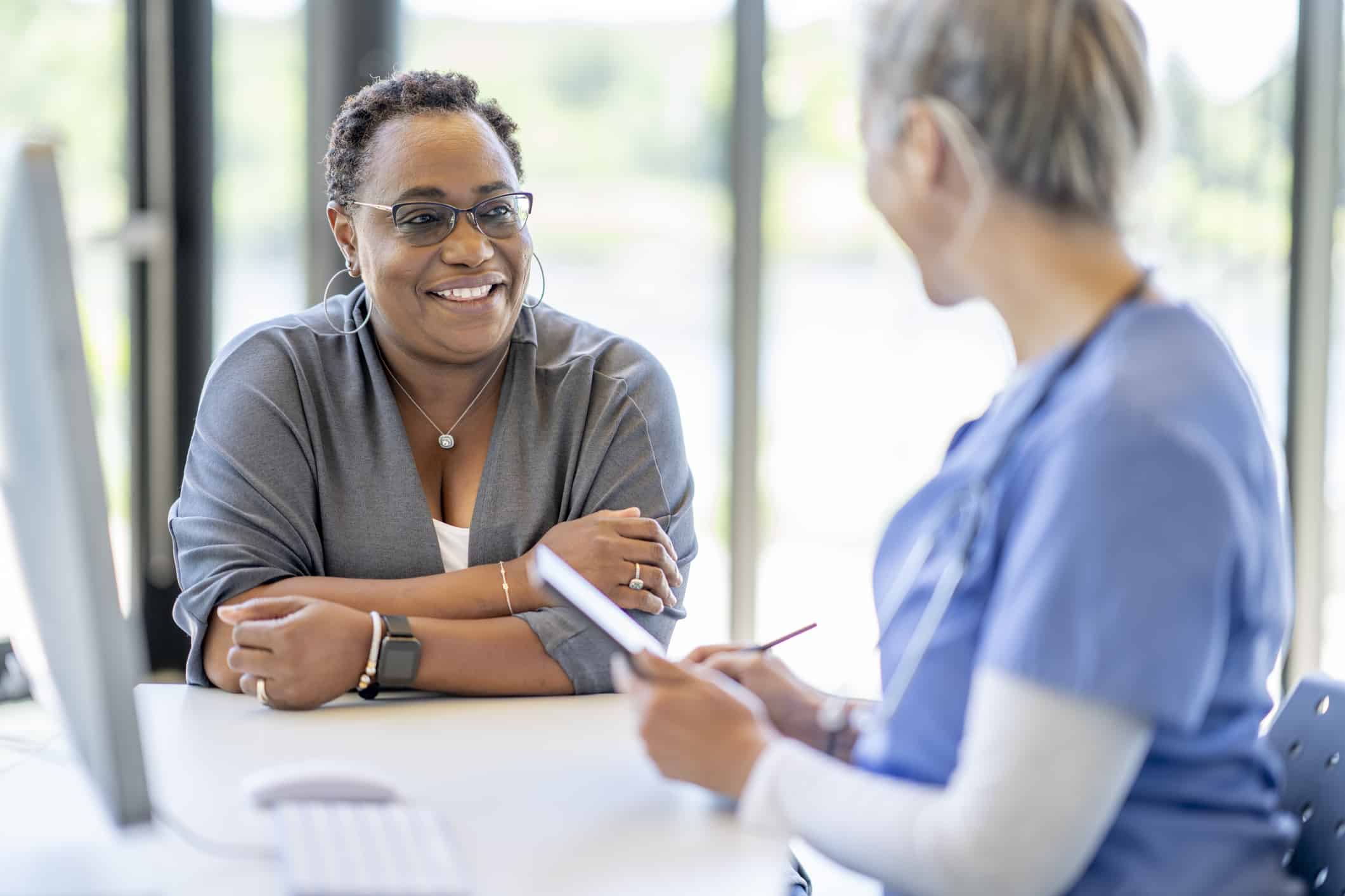 A middle aged woman of African decent, sits with her doctor at her desk as they meet to discuss her health.  The woman is dressed casually and smiling at the doctor as she listens attentively.