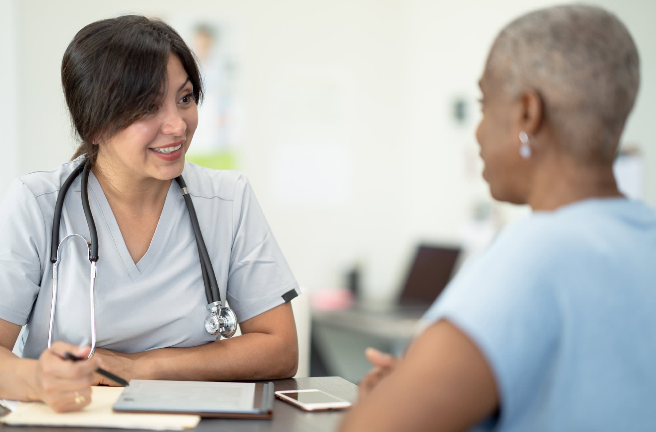 Making a Health Plan A female doctor sits down with a senior patient at her desk as they discuss and come up with a plan for her health care needs. She is wearing white scrubs and has a stethoscope around her neck as she reviews the patients file and recent test results on her tablet out in front of them.
