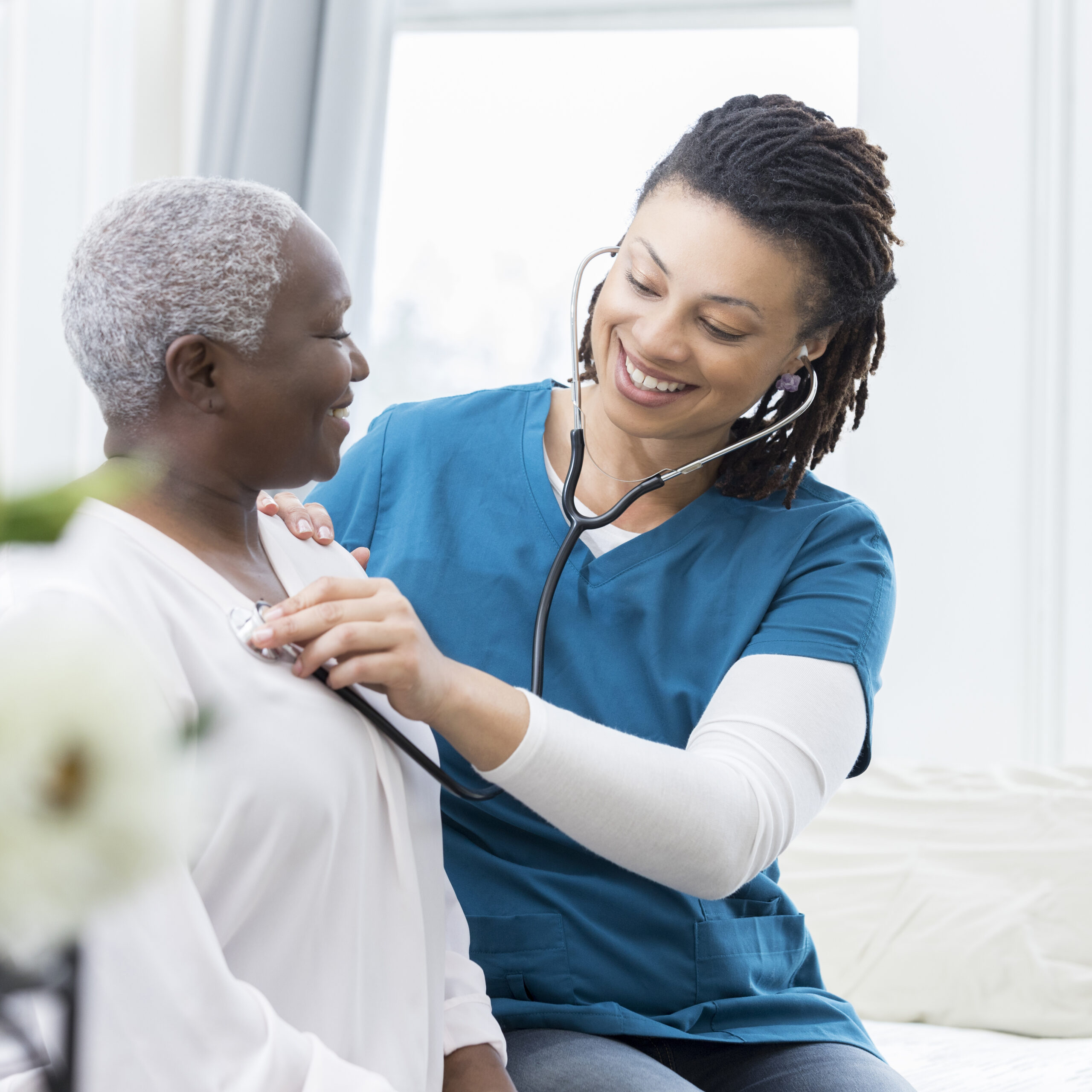 Cheerful young female nurse smiles while checking a senior adult's heartbeat.