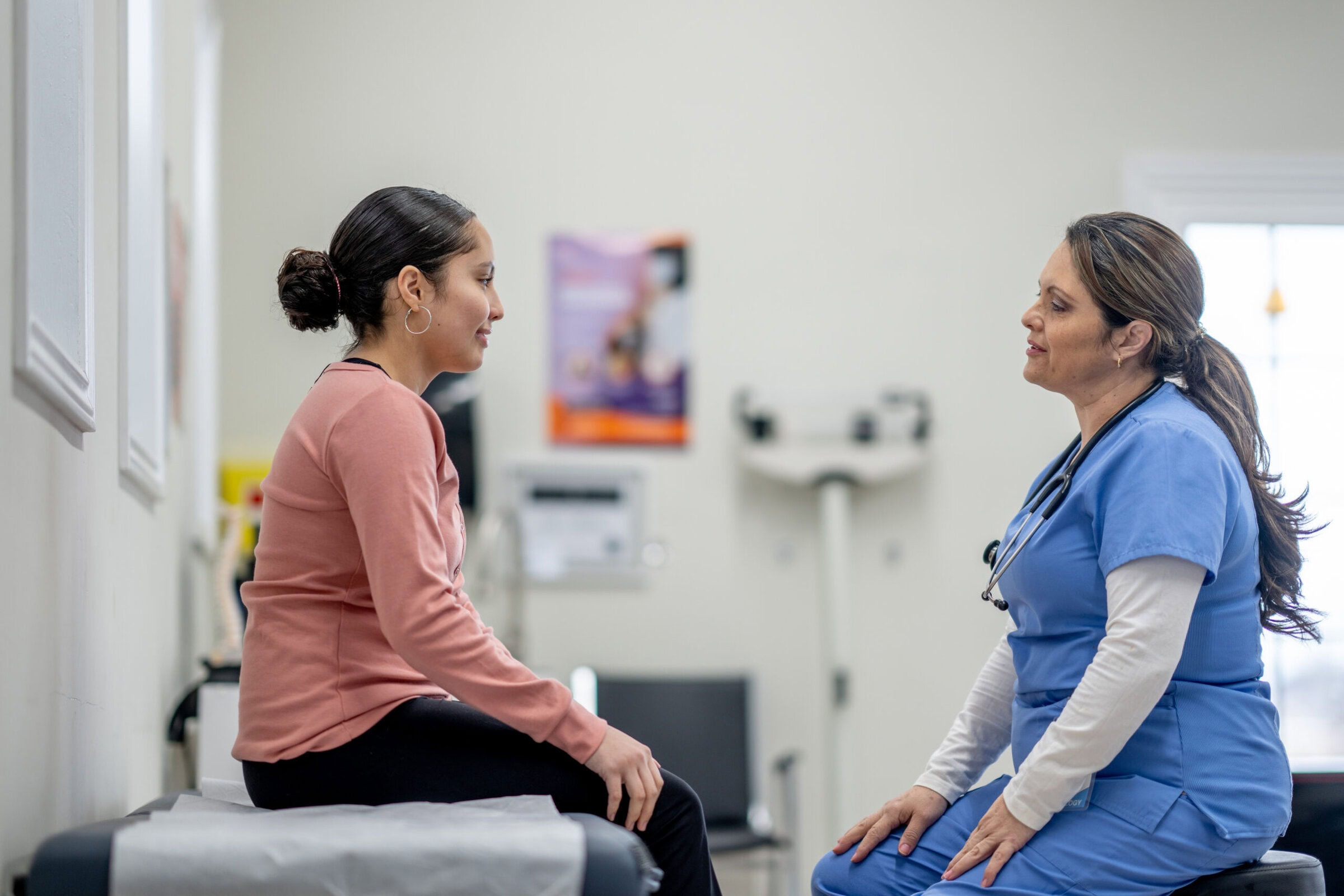 A female sits up on an exam table during a routine check-up.  She is dressed casually and her female health care provider is of Hispanic decent, is seated in front of her as they talk.